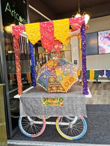 Decorated cart with coloured emroided fabric umbrella. Each of the four posts wrapped with coloured patterned fabric from India. The table cloth has Diwali in coloured fabric letters in English and Hindi. The bike wheels of the court, are pink, blue and red pastel colours. It's bright and happy looking cart.